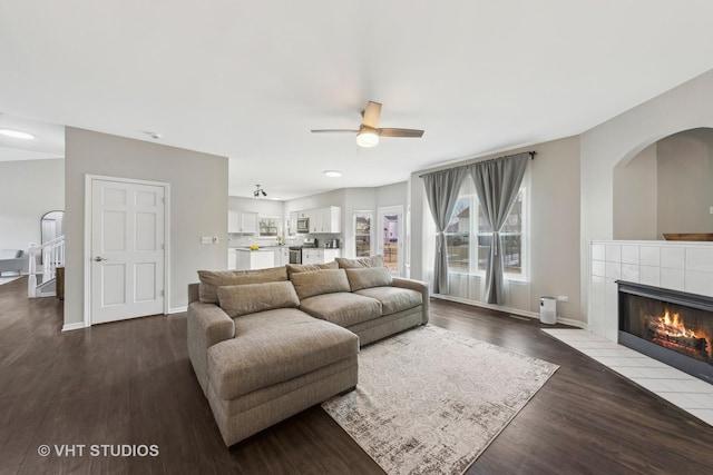 living room with a ceiling fan, baseboards, a tiled fireplace, and wood finished floors