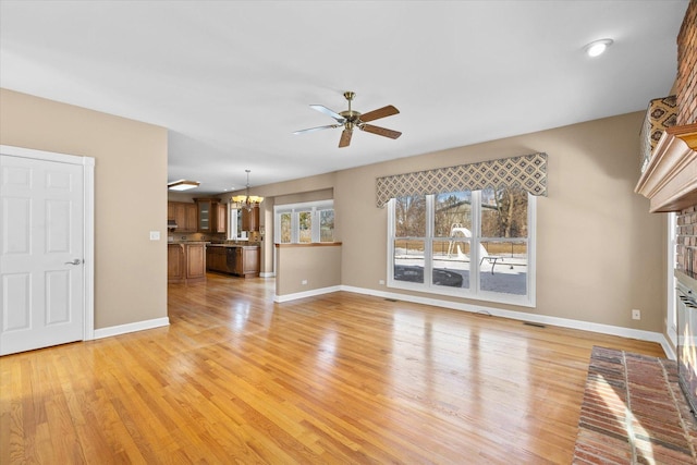 unfurnished living room featuring ceiling fan with notable chandelier, visible vents, baseboards, light wood-type flooring, and a brick fireplace