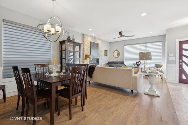 dining space with a stone fireplace, light wood-style flooring, ceiling fan with notable chandelier, and recessed lighting