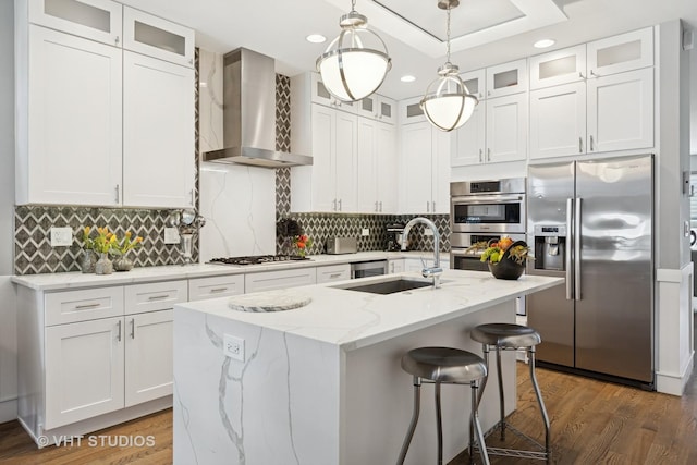 kitchen with an island with sink, appliances with stainless steel finishes, wood finished floors, wall chimney exhaust hood, and a sink