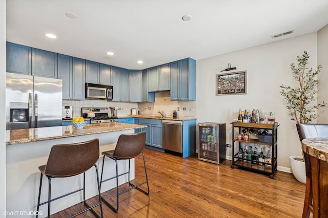 kitchen with beverage cooler, wood finished floors, stainless steel appliances, blue cabinetry, and backsplash