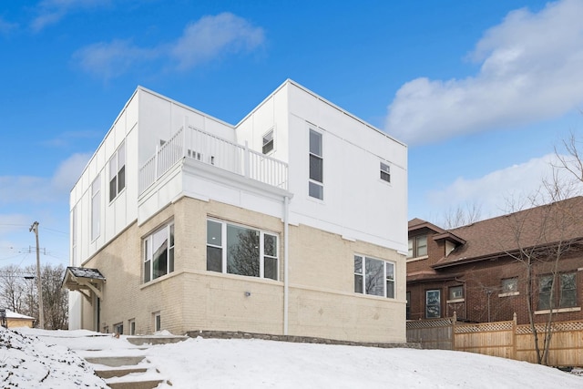 snow covered rear of property featuring brick siding, fence, and a balcony