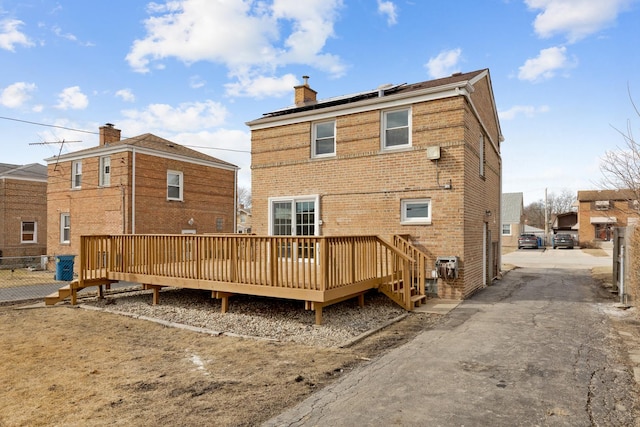 rear view of house with brick siding, a chimney, roof mounted solar panels, fence, and a deck