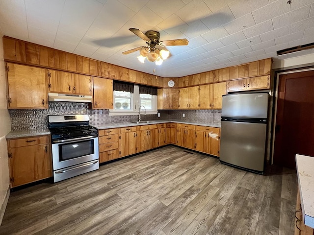 kitchen with under cabinet range hood, wood finished floors, a sink, appliances with stainless steel finishes, and brown cabinets