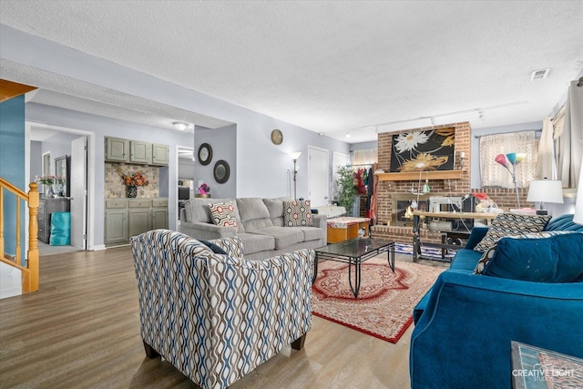 living room featuring a textured ceiling, wood finished floors, visible vents, stairway, and a brick fireplace