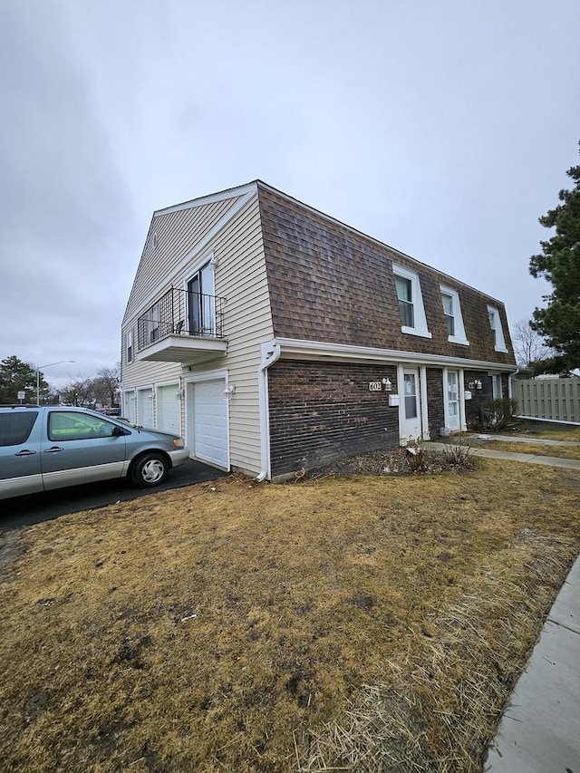 view of side of property with a garage, mansard roof, a balcony, roof with shingles, and brick siding