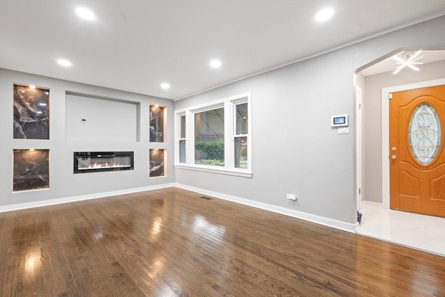 unfurnished living room featuring arched walkways, recessed lighting, a glass covered fireplace, wood finished floors, and baseboards