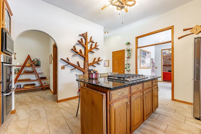 kitchen with arched walkways, a center island, stainless steel appliances, brown cabinetry, and dark stone counters