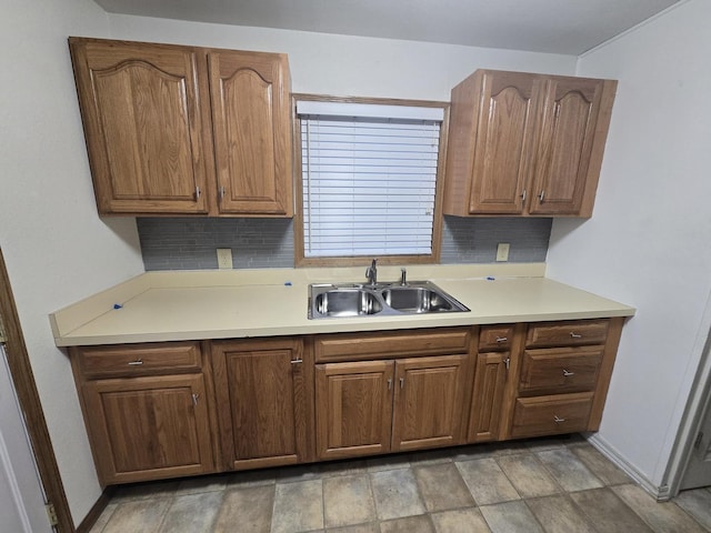 kitchen featuring light countertops, backsplash, brown cabinetry, a sink, and baseboards