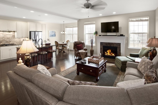 living area featuring a premium fireplace, ceiling fan with notable chandelier, dark wood finished floors, and recessed lighting