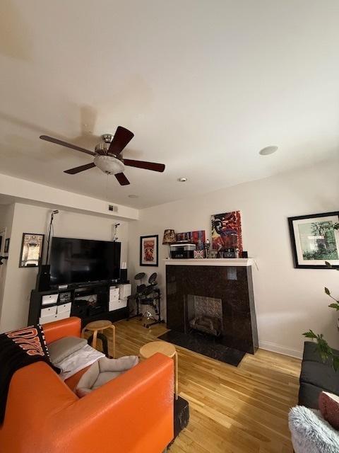 living area featuring a fireplace with flush hearth, a ceiling fan, baseboards, and wood finished floors