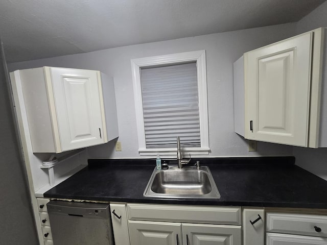 kitchen featuring stainless steel dishwasher, dark countertops, a sink, and white cabinetry