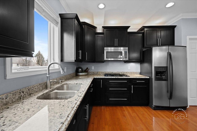 kitchen with appliances with stainless steel finishes, dark cabinets, light stone countertops, crown molding, and a sink