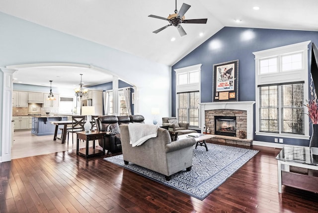 living room with baseboards, hardwood / wood-style flooring, a stone fireplace, high vaulted ceiling, and ceiling fan with notable chandelier