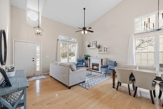 living room featuring light wood-style floors, a glass covered fireplace, high vaulted ceiling, baseboards, and ceiling fan with notable chandelier