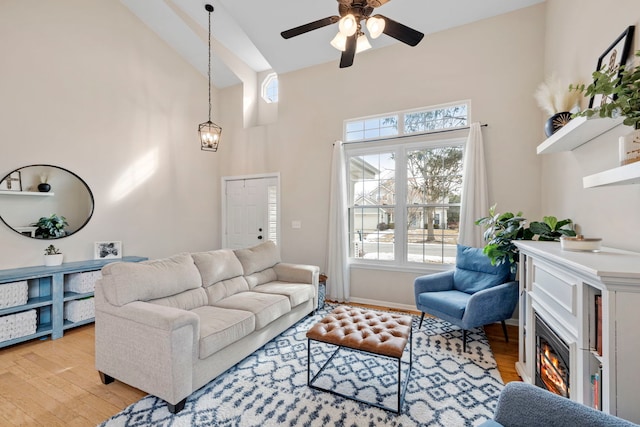 living room with light wood-type flooring, ceiling fan with notable chandelier, high vaulted ceiling, and a glass covered fireplace