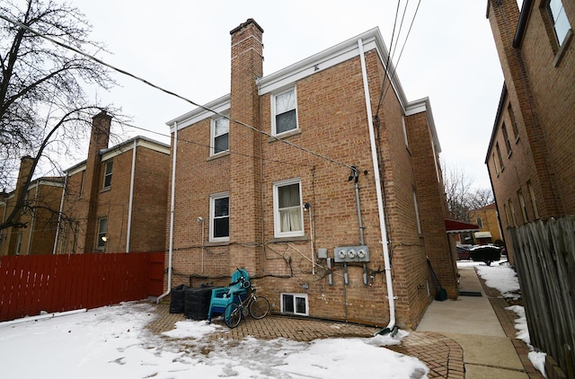 snow covered rear of property with a chimney, fence, and brick siding