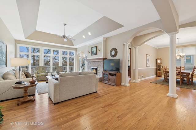 living room with a brick fireplace, a raised ceiling, a ceiling fan, light wood-style floors, and ornate columns