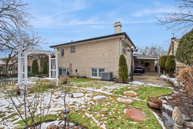 snow covered house with brick siding, a chimney, central AC unit, and a pergola