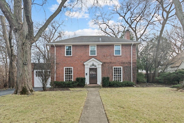 colonial-style house featuring brick siding, a chimney, a front yard, a garage, and driveway