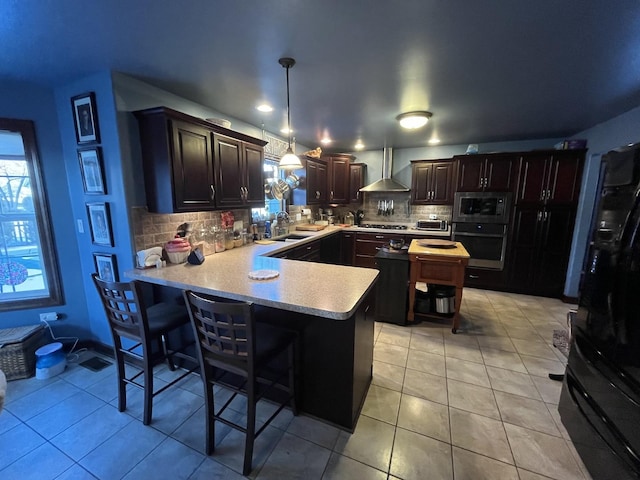 kitchen with a peninsula, stainless steel appliances, light countertops, wall chimney range hood, and hanging light fixtures