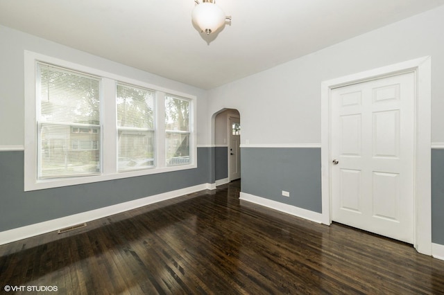 empty room with arched walkways, dark wood-style floors, visible vents, and baseboards