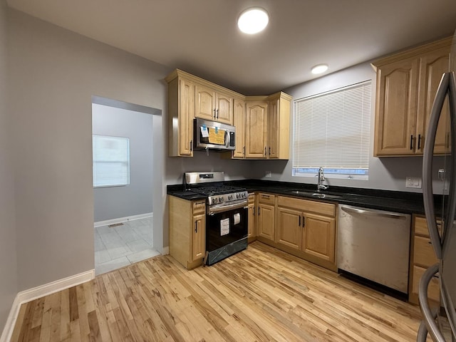 kitchen with appliances with stainless steel finishes, dark stone counters, a sink, light wood-type flooring, and baseboards