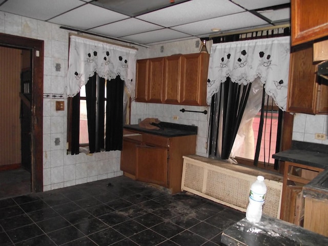 kitchen with a drop ceiling, dark tile patterned floors, tile walls, brown cabinetry, and dark countertops