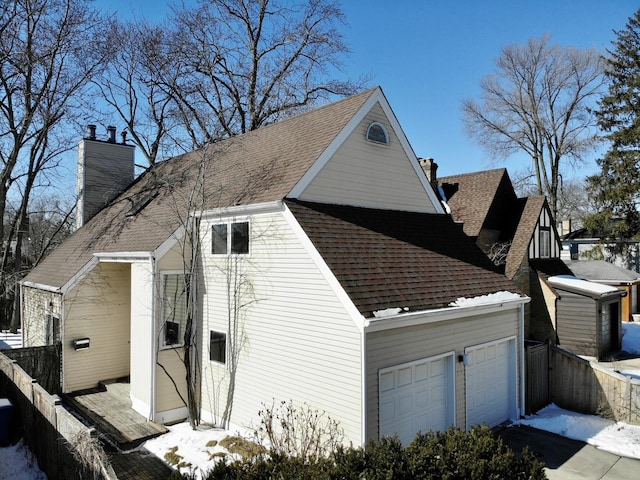 view of snow covered exterior featuring a garage, a shingled roof, a chimney, and fence