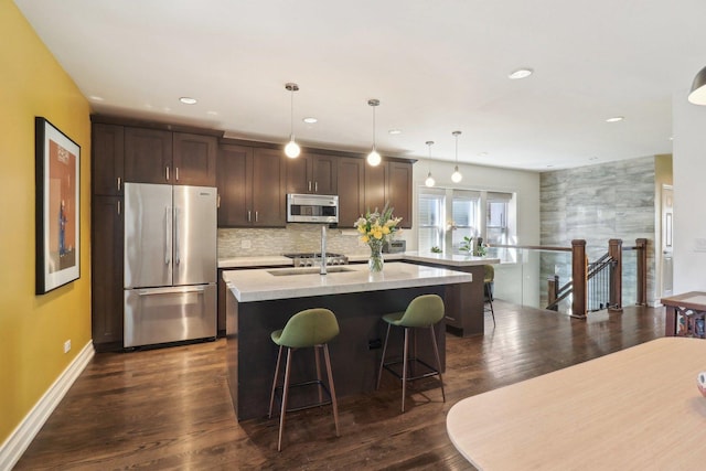 kitchen featuring a kitchen island with sink, stainless steel appliances, dark wood-style flooring, dark brown cabinets, and a kitchen bar