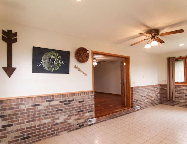 spare room featuring light tile patterned floors, a wainscoted wall, brick wall, visible vents, and a ceiling fan