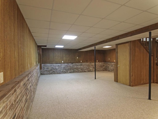 finished below grade area with a paneled ceiling, light colored carpet, wood walls, and brick wall