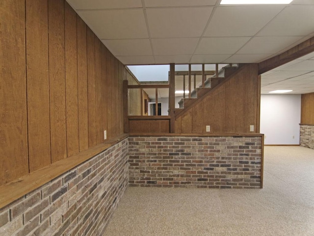 finished below grade area featuring a paneled ceiling, brick wall, wood walls, carpet flooring, and stairway