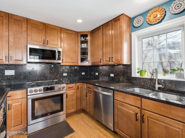 kitchen with appliances with stainless steel finishes, brown cabinetry, glass insert cabinets, a sink, and dark stone counters