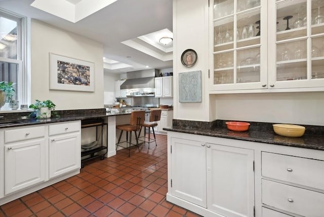 kitchen with glass insert cabinets, a raised ceiling, white cabinetry, and wall chimney range hood