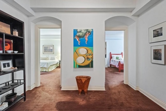 hallway featuring built in shelves, arched walkways, baseboards, and dark colored carpet