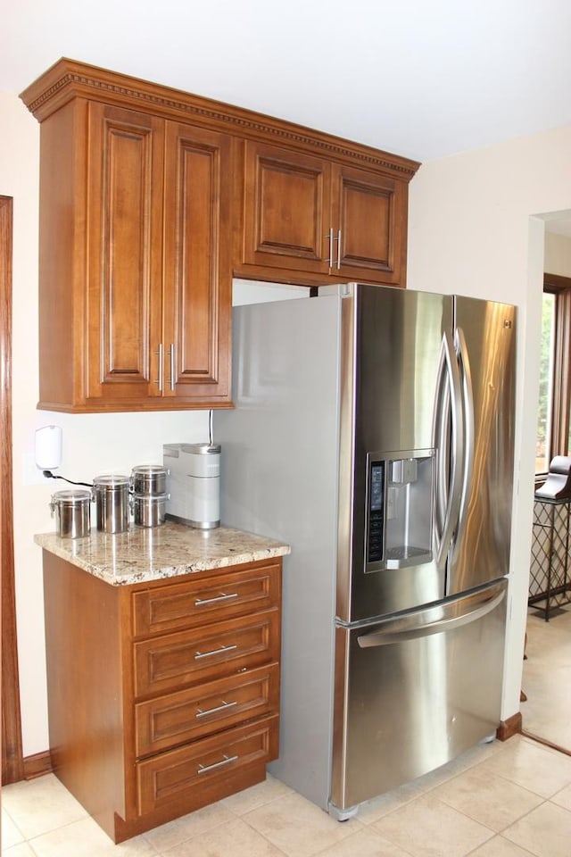 kitchen with brown cabinetry, light tile patterned flooring, stainless steel fridge with ice dispenser, and light stone countertops