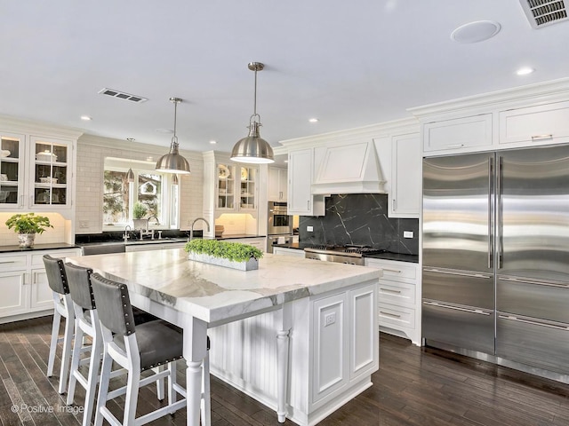 kitchen featuring premium range hood, a kitchen island, visible vents, appliances with stainless steel finishes, and glass insert cabinets