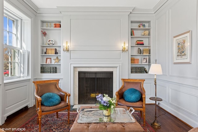 sitting room featuring built in features, a decorative wall, dark wood-type flooring, a fireplace with flush hearth, and ornamental molding