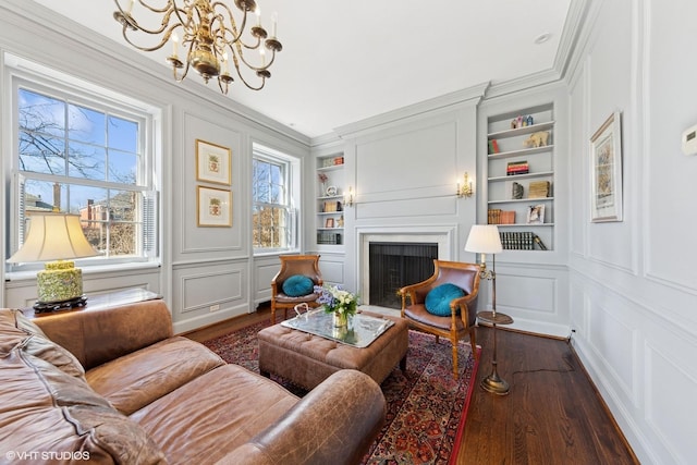 living area with built in features, dark wood-style flooring, a decorative wall, a fireplace with flush hearth, and ornamental molding