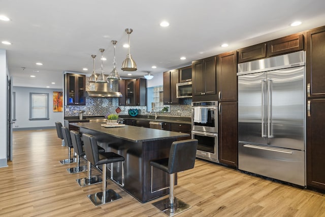 kitchen featuring stainless steel appliances, dark countertops, a kitchen bar, light wood-style floors, and wall chimney exhaust hood