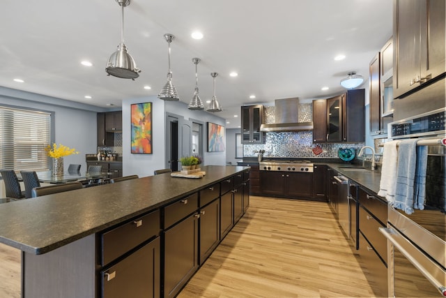 kitchen with dark countertops, a sink, a kitchen island, light wood-type flooring, and wall chimney exhaust hood