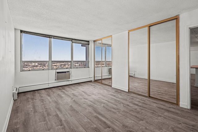 unfurnished bedroom with a textured ceiling, a baseboard heating unit, wood finished floors, a wall mounted AC, and two closets