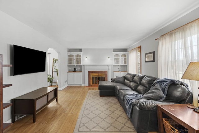 living room featuring arched walkways, light wood finished floors, baseboards, and a brick fireplace