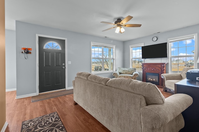 living area with a glass covered fireplace, ceiling fan, baseboards, and wood finished floors