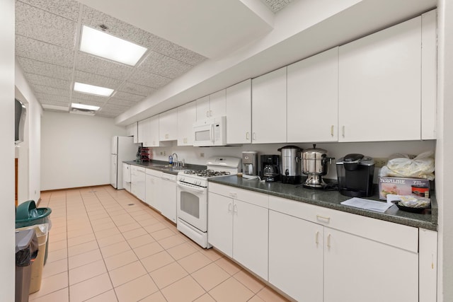 kitchen with white appliances, dark countertops, a sink, a drop ceiling, and white cabinetry