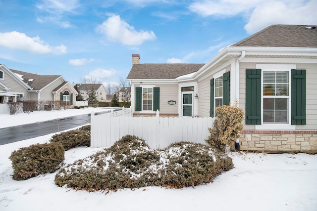 view of snow covered exterior featuring a shingled roof, stone siding, fence, and a chimney