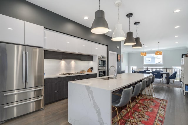 kitchen with an island with sink, dark wood-style floors, appliances with stainless steel finishes, decorative light fixtures, and white cabinetry