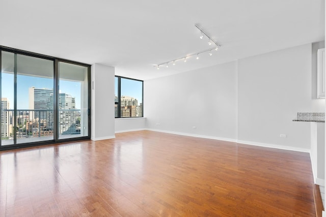 unfurnished room featuring wood finished floors, baseboards, a view of city, a wall of windows, and rail lighting