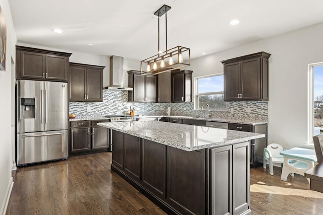 kitchen with a center island, hanging light fixtures, light stone countertops, wall chimney exhaust hood, and stainless steel fridge